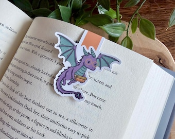 May include: A whimsical bookmark featuring a purple dragon with glasses and teal wings, holding books. The dragon is placed on the pages of an open book, with text visible. The bookmark has an orange tab.
