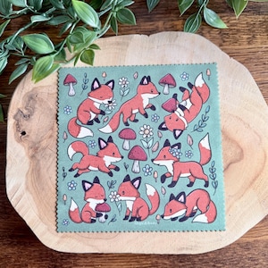 May include: A square, green cloth with a repeating pattern of cartoon foxes, mushrooms, and flowers. The foxes are orange with white and black accents. The mushrooms are red with white spots. The flowers are white.
