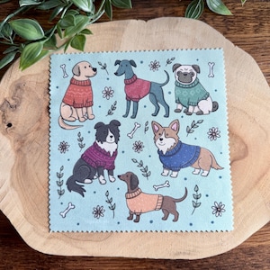 Può includere: Un quadrato azzurro con illustrazioni di sei diverse razze di cani che indossano maglioni. I cani sono circondati da ossa e motivi floreali. I cani sono di vari colori, tra cui beige, nero e marrone.