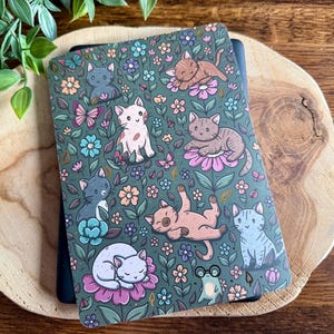 以下が含まれることがあります： 漫画の猫、花、蝶の気まぐれなパターンが特徴の、ティール色のノートカバー。 猫はさまざまなポーズをとっており、デザインにはメガネをかけた小さなカエルが含まれています。 ノートは木製の表面にあります。