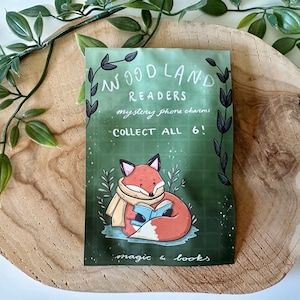 Puede incluir: Un paquete verde con el texto "Woodland Readers" y un zorro de dibujos animados leyendo un libro. El paquete también dice "¡Colecciona los 6!" y "magic & books". El paquete está sobre una superficie de madera con hojas verdes.