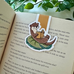 Peut inclure: Une loutre brune et beige lisant un livre assise sur un nénuphar vert. La loutre porte une chemise marron clair et a le ventre blanc. Le livre est brun avec une tranche jaune. L'arrière-plan est une image floue d'un livre.