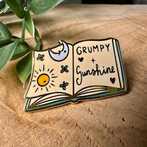 Può includere: Spilla in smalto dorata a forma di libro aperto con un sole, una luna e cuori sulle pagine. Il testo sul libro recita "Grumpy + Sunshine".