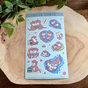 Peut inclure: Une planche d'autocollants intitulée "OTTER STORIES" présente des illustrations d'otaries lisant des livres, se câlinant et flottant dans l'eau. Les loutres sont de couleur marron et rose, avec des détails bleus et blancs. La planche est posée sur une surface en bois.