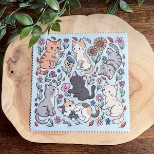 May include: A light blue square fabric with a charming illustration of cartoon cats and flowers. The cats are depicted in different poses, with colours including orange, grey, black, and white. The fabric is displayed on a wooden surface.