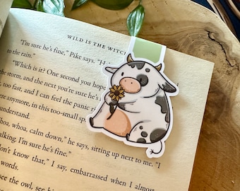 May include: A white and black cow bookmark with a sunflower in its mouth. The bookmark is on a book with the text "WILD IS THE WITCI" and "I'm sure he's fine," Pike says. "H" visible.