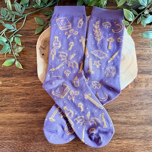 May include: A pair of purple crew socks featuring a repeating pattern of yellow line-art illustrations including a dragon, a sword, a book, a candle, a key, a crystal ball, a crescent moon, a star, and various small botanical elements.