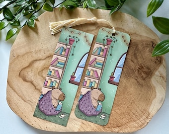 May include: Two illustrated bookmarks featuring a capybara reading a book in a cozy room with a bookshelf, a window, and a potted plant. The bookmarks are printed on cardstock and have a tassel.
