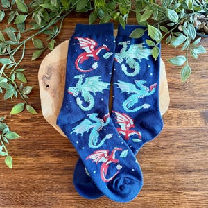 Könnte beinhalten: Ein Paar marineblaue Socken mit einem wiederkehrenden Muster aus kleinen, stilisierten Drachen in Rot- und Hellblautönen. Die Drachen sind in verschiedenen Flugposen dargestellt, durchsetzt mit winzigen weißen Punkten, die wie Sterne wirken.