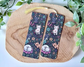 May include: Two decorative bookmarks with a cat theme. The bookmarks feature a dark green background with illustrations of cats, flowers, and butterflies. One bookmark has a tassel. The bookmarks are on a wooden surface.