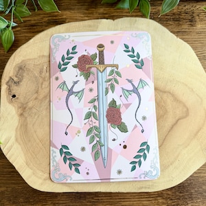 Könnte beinhalten: Eine rosa-weiße Tarotkarte mit einem Schwert, Rosen und zwei Drachen. Das Schwert hat einen goldenen Knauf und ist von grünen Blättern umgeben. Die Karte liegt auf einer Holzoberfläche.
