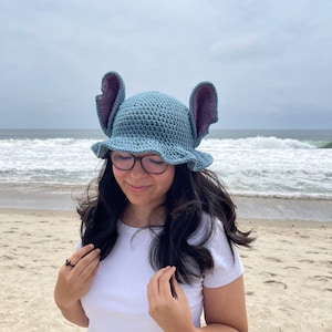 May include: A blue crochet hat with large, upright ears and a ruffled brim. The hat's interior is a contrasting purple colour. The hat is designed to resemble a cartoon character. The hat is worn by a person on a beach.