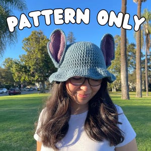 Pode incluir: Um chapéu de crochê azul-acinzentado com orelhas grandes e verticais em um tom roxo contrastante. O chapéu tem uma copa arredondada e uma aba ligeiramente alargada. O texto "PATTERN ONLY" é exibido em letras brancas.