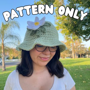 Op de afbeelding: Een lichtgroene gehaakte vissershoed met een witte en gele bloem en groene bladeren. De hoed heeft een brede rand. De tekst "PATTERN ONLY" staat in het wit, omlijnd met zwart, bovenaan de afbeelding.