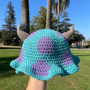 Può includere: Un cappello a secchiello all'uncinetto con macchie viola e blu e due corna marroni. Il cappello è tenuto sollevato in aria contro uno sfondo di erba verde.