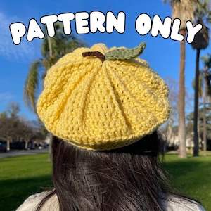 Pode incluir: Um gorro de crochê amarelo com um detalhe de folha verde no topo. O gorro é usado por uma pessoa com cabelo comprido e escuro.
