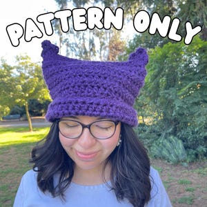 May include: A purple crocheted hat with pointed ears, worn by a person wearing glasses. The text "PATTERN ONLY" is displayed in a bubble font above the hat. The hat is a vibrant shade of purple.