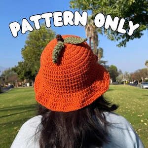 May include: An orange crochet bucket hat with a green leaf and stem detail. The hat has the text "PATTERN ONLY" in white. The hat is a bright orange color and appears to be handmade. The background is a sunny outdoor scene.