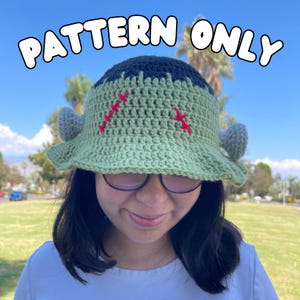 May include: A handmade crochet bucket hat in a Frankenstein monster design. The hat features a green brim, a dark blue top, and gray side pieces. Red stitches are embroidered on the front. The text "PATTERN ONLY" is displayed above the hat.