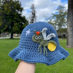 May include: A blue crocheted bucket hat with a green and yellow firefly design. The firefly has googly eyes and a white star is embroidered on the hat.