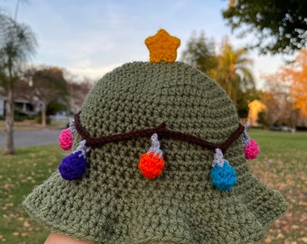 Peut inclure: Chapeau bob crocheté vert en forme de sapin de Noël avec une étoile jaune sur le dessus et des lumières colorées autour du bord.