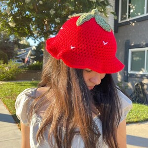 May include: A red crocheted strawberry bucket hat with green leaves on top. The hat has white stitching details.
