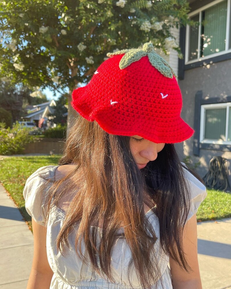 May include: A red crocheted strawberry bucket hat with green leaves on top. The hat has white stitching details.