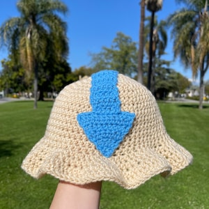 Puede incluir: Un sombrero de pescador de ganchillo beige con un emblema de flecha azul. El sombrero tiene un borde con volantes y está hecho de hilo de color claro. La flecha azul está centrada en la parte delantera del sombrero.