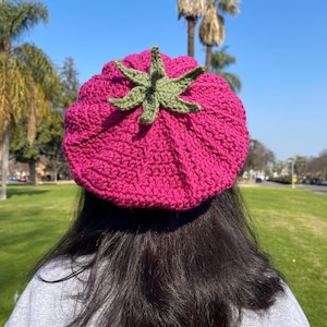May include: A pink crocheted beret with a green crocheted leaf design on top. The beret is worn by a person with long dark hair.
