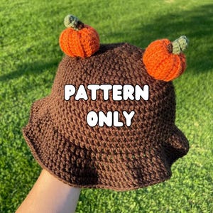 May include: A brown crochet bucket hat with two orange crocheted pumpkins attached to the brim. The hat has a "PATTERN ONLY" text overlay.