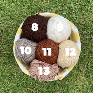 Puede incluir: Una cesta llena de ovillos de lana de varios colores, incluyendo marr&oacute;n, crema, beige y gris. La cesta tiene un forro a cuadros amarillos y blancos. Los ovillos de lana est&aacute;n numerados 8, 9, 10, 11, 12 y 13.
