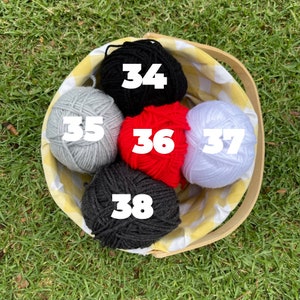 Puede incluir: Una cesta de picnic llena de seis ovillos de lana de varios colores: negro, gris, rojo y blanco. La cesta tiene un forro a cuadros amarillos y blancos y un asa de madera. Los n&uacute;meros 34, 35, 36, 37 y 38 son visibles en la lana.