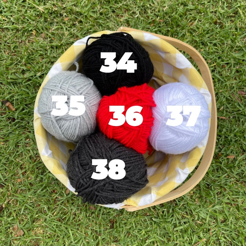 Puede incluir: Una cesta de picnic llena de seis ovillos de lana de varios colores: negro, gris, rojo y blanco. La cesta tiene un forro a cuadros amarillos y blancos y un asa de madera. Los n&uacute;meros 34, 35, 36, 37 y 38 son visibles en la lana.