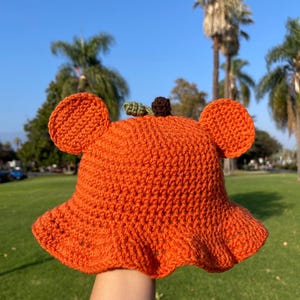 Puede incluir: Sombrero de cubo de punto de ganchillo naranja con un tallo verde y una parte superior marrón que se asemeja a una calabaza. El sombrero tiene dos orejas caídas a los lados.