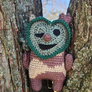Könnte beinhalten: Ein handgefertigtes Häkeltier mit herzförmigem Gesicht und zwinkerndem Auge, eingebettet in einem Baum. Das Tier hat einen grünen Rand um das Gesicht, einen braunen Körper mit cremefarbenen Streifen und kleine Arme und Beine.
