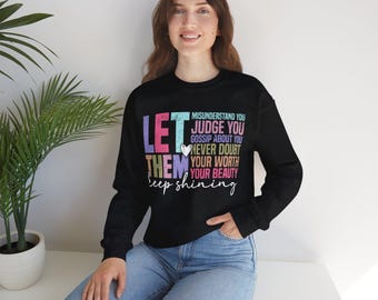 Op de afbeelding: Zwart sweatshirt met een kleurrijke afbeelding met de tekst "Laat ze je misverstaan, beoordelen, roddelen over je, twijfel nooit aan je waarde, je schoonheid, blijf stralen".