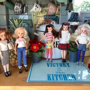 May include: Five dolls dressed in different outfits stand on a wooden surface. The dolls are all wearing different styles of clothing, including a plaid skirt, a striped shirt, and a white sweater. The dolls are standing in front of a blue and white sign that says "Victory is in the kitchen".