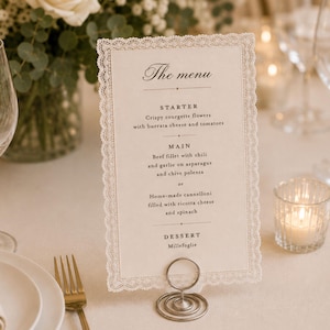 May include: A rectangular menu card with a delicate white lace border, held upright by a silver spiral wire stand. The card features black printed text listing courses including starter, main, and dessert, set against a cream-colored background.