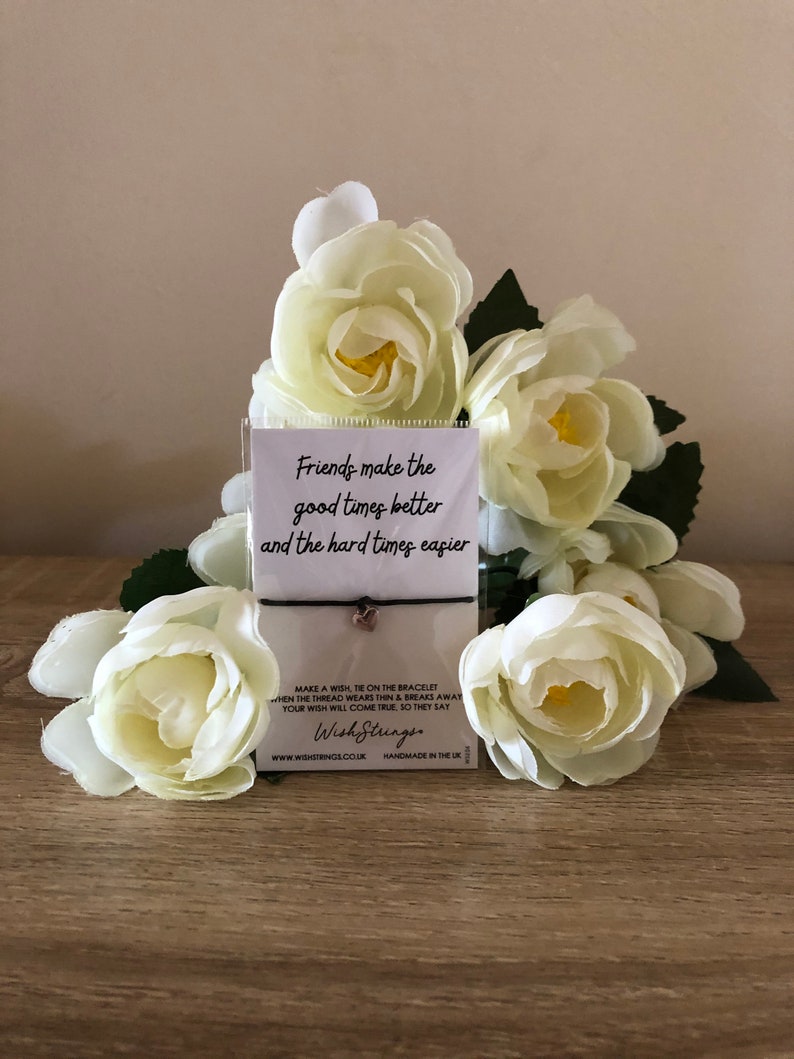 Op de afbeelding: Een zwart koordarmband met een gouden bedel waarop staat "Friends make the good times better and the hard times easier". De armband is verpakt op een witte kaart met het WishStrings-logo en websiteadres.