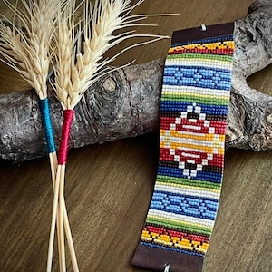 Puede incluir: Un brazalete de cuentas coloridas con un patrón geométrico en tonos de rojo, amarillo, azul y verde. El brazalete está hecho con cuentas pequeñas y estrechamente tejidas y tiene un respaldo de cuero marrón con un cierre plateado.