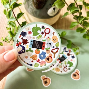 Puede incluir: Una pegatina redonda con un borde verde claro que presenta ilustraciones coloridas del videojuego Animal Crossing. La pegatina incluye una consola Switch, cerezas y una bolsa de dinero.