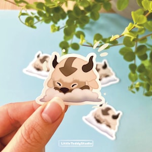 May include: A sticker featuring a cartoon bison-like creature with brown and tan fur, black horns, and a brown arrow marking on its head. The creature is resting on a cloud. The sticker is held by a person's hand. The text "Little Teddy Studio" is at the bottom.