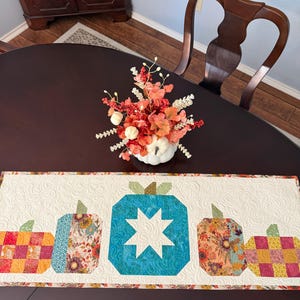 May include: A rectangular quilted table runner featuring a white background with a swirling stitch pattern. The runner displays five fabric pumpkins in a row, showcasing various patterns including teal with a white star, floral, and checkered prints.