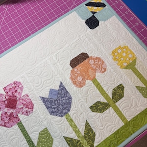 May include: A quilted floral wall hanging featuring a bee and four stylized flowers in shades of pink, orange, purple, and yellow. The flowers have green stems and leaves, set against a white background with a quilted pattern. The border is light blue.
