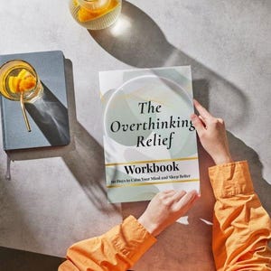 May include: A workbook titled "The Overthinking Relief Workbook" with a light green and white cover. The book is held by a person wearing an orange shirt. A blue notebook and a glass of orange-infused beverage are also visible.