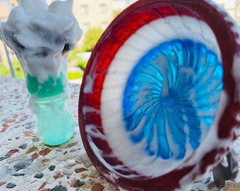 May include: A red, white, and blue bowl with a blue swirl design is covered in white shaving cream. A green and white shaving brush is also covered in white shaving cream.