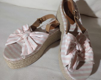 May include: A pair of pink and white striped wedge sandals with a bow detail on the front. The sandals have a brown leather strap and a jute wedge sole.