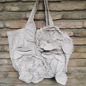 May include: A light grey canvas tote bag with a large, three-dimensional flower design. The flower is made of multiple layers of fabric, giving it a textured look.