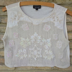 Könnte beinhalten: Cremefarbenes ärmelloses Oberteil mit gehäkeltem und gesticktem Blumendesign. Das Oberteil hat einen runden Ausschnitt und eine verkürzte Länge. Das florale Design umfasst Blatt- und Blumenmuster mit dezenten Pastellakzenten. Das Markenetikett "TOPSHOP" ist sichtbar.