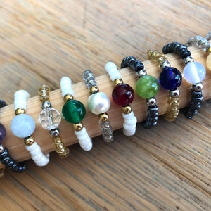Può includere: Una collezione di anelli di perline con vari accenti di pietre preziose e cristalli. Ogni anello presenta una perla centrale in colori come viola, blu, verde e trasparente, su una fascia di perline più piccole. Distanziatori dorati e argentati aggiungono dettagli.